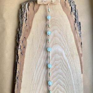 May include: A rustic wooden wall hanging featuring a natural wood slab with a bark edge. A string of light blue glass beads and small green beads hangs from a small wooden dowel at the top, creating a decorative element.