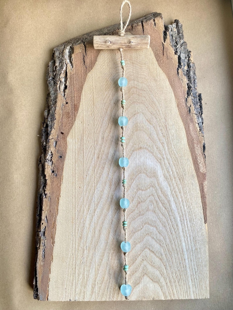 May include: A rustic wooden wall hanging featuring a natural wood slab with a bark edge. A string of light blue glass beads and small green beads hangs from a small wooden dowel at the top, creating a decorative element.
