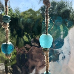 May include: Close-up of a decorative hanging ornament featuring light blue glass beads and smaller, darker blue beads strung on natural twine. The beads have a sea glass appearance. The background is a blurred view of trees and sky.