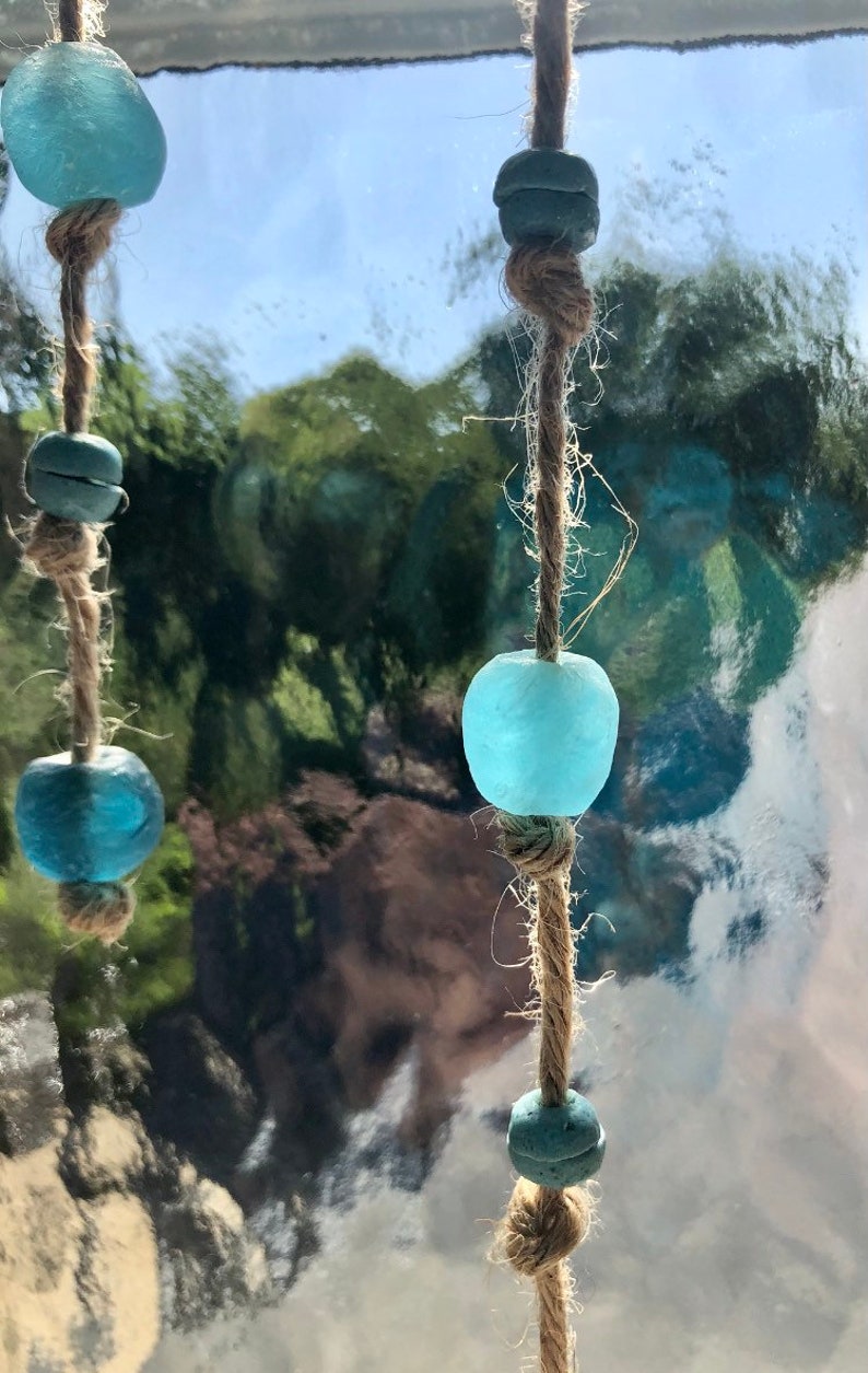 May include: Close-up of a decorative hanging ornament featuring light blue glass beads and smaller, darker blue beads strung on natural twine. The beads have a sea glass appearance. The background is a blurred view of trees and sky.