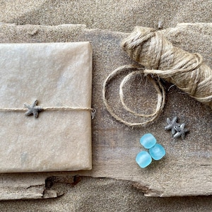 Pode incluir: Um presente embrulhado em papel castanho, amarrado com barbante e um pingente de estrela do mar. A imagem tamb&eacute;m inclui um rolo de barbante, dois pingentes de estrela do mar e tr&ecirc;s peda&ccedil;os de vidro do mar azul claro. Os itens est&atilde;o em uma superf&iacute;cie de madeira com areia.