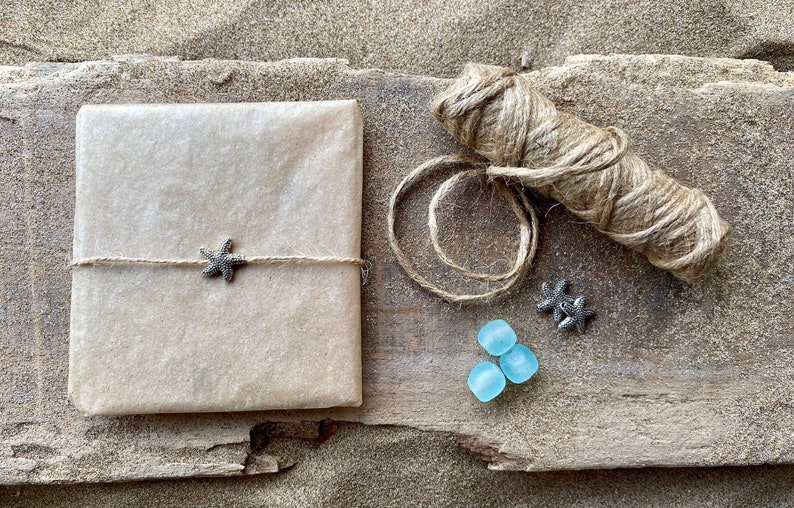 Pode incluir: Um presente embrulhado em papel castanho, amarrado com barbante e um pingente de estrela do mar. A imagem tamb&eacute;m inclui um rolo de barbante, dois pingentes de estrela do mar e tr&ecirc;s peda&ccedil;os de vidro do mar azul claro. Os itens est&atilde;o em uma superf&iacute;cie de madeira com areia.