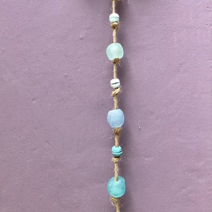 May include: A handcrafted wall hanging featuring a natural wood piece and a strand of sea glass beads in shades of blue and white. The beads are strung on a natural twine, creating a coastal or bohemian aesthetic.