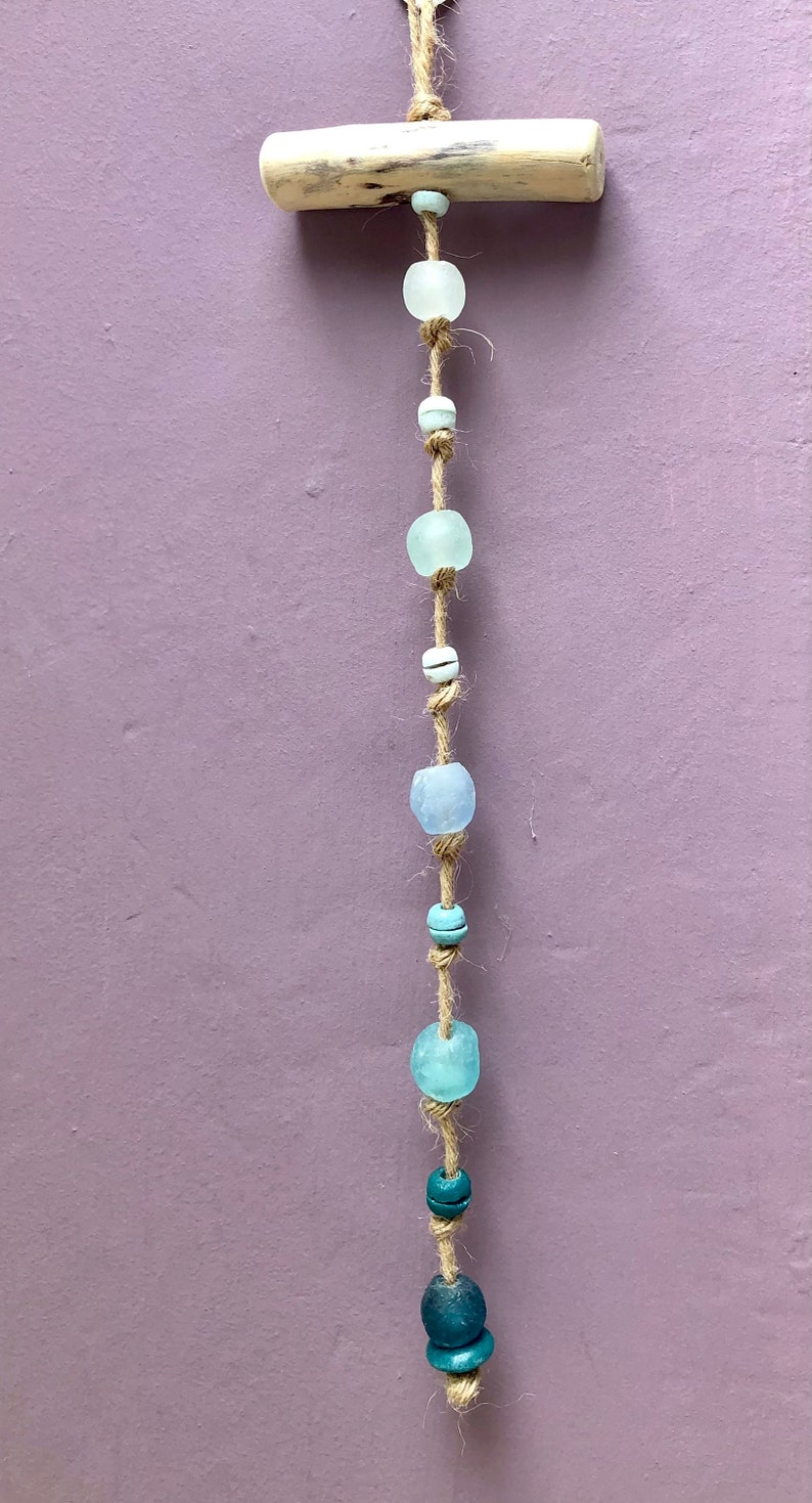 May include: A handcrafted wall hanging featuring a natural wood piece and a strand of sea glass beads in shades of blue and white. The beads are strung on a natural twine, creating a coastal or bohemian aesthetic.