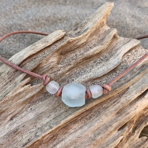 May include: A simple necklace made with brown leather cord and three white sea glass beads. The necklace is displayed on a piece of driftwood.