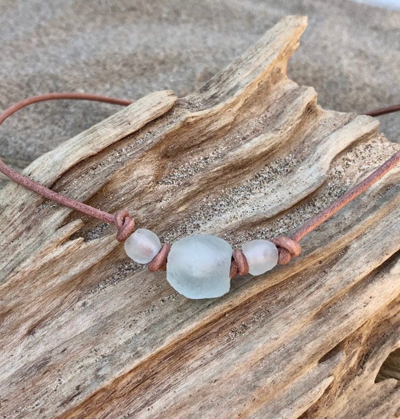 May include: A simple necklace made with brown leather cord and three white sea glass beads. The necklace is displayed on a piece of driftwood.