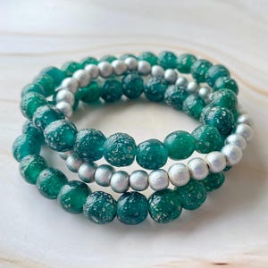 May include: Three beaded bracelets. Two bracelets feature large, round, teal-green beads with a speckled texture. The third bracelet has smaller, silver-coloured beads. The bracelets are stacked together.