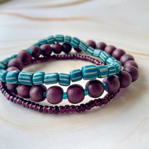 May include: Three stacked bracelets. The top bracelet features turquoise and white striped beads. The middle bracelet has dark purple wooden beads. The bottom bracelet is made of small, dark purple beads. All bracelets have turquoise accents.