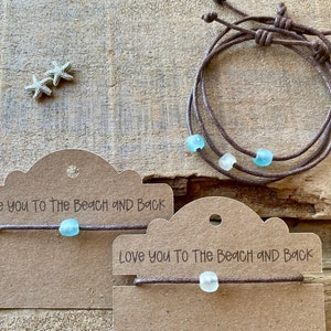Pode incluir: Pulseiras de cord&atilde;o marrom com contas de vidro do mar. Uma pulseira tem tr&ecirc;s fios, as outras t&ecirc;m um. As pulseiras s&atilde;o exibidas em cart&otilde;es marrons com o texto "Love you to The Beach and Back". Duas pequenas estrelas do mar tamb&eacute;m s&atilde;o vis&iacute;veis.