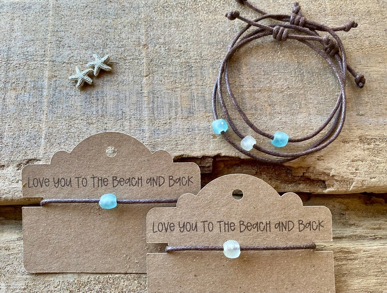 Pode incluir: Pulseiras de cord&atilde;o marrom com contas de vidro do mar. Uma pulseira tem tr&ecirc;s fios, as outras t&ecirc;m um. As pulseiras s&atilde;o exibidas em cart&otilde;es marrons com o texto "Love you to The Beach and Back". Duas pequenas estrelas do mar tamb&eacute;m s&atilde;o vis&iacute;veis.