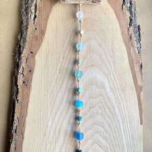 May include: A rustic wall hanging featuring a wooden plank with a natural bark edge. A string of blue and clear glass beads hangs from a small wooden bar at the top. The beads vary in size and shade, creating a gradient effect.