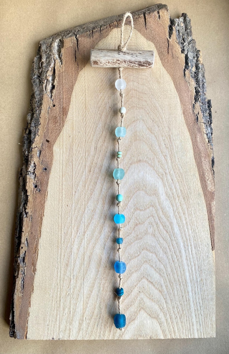 May include: A rustic wall hanging featuring a wooden plank with a natural bark edge. A string of blue and clear glass beads hangs from a small wooden bar at the top. The beads vary in size and shade, creating a gradient effect.