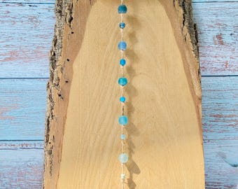 May include: A vertical wall hanging featuring a horizontal piece of light-colored driftwood suspended by a jute cord. A single strand of various blue and clear glass beads in different sizes and shapes hangs from the center of the driftwood piece.