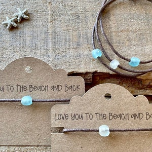 Pode incluir: Pulseiras de cord&atilde;o marrom com contas de vidro do mar. As pulseiras s&atilde;o exibidas com pequenas estrelas do mar prateadas e etiquetas que dizem "Love you to the beach and back."