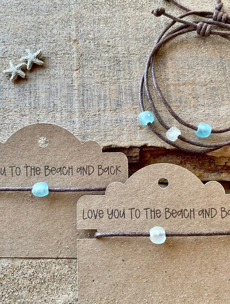 Pode incluir: Pulseiras de cord&atilde;o marrom com contas de vidro do mar. As pulseiras s&atilde;o exibidas com pequenas estrelas do mar prateadas e etiquetas que dizem "Love you to the beach and back."