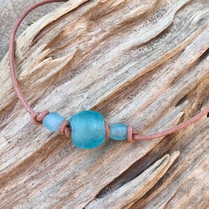 May include: A light blue glass bead necklace with a brown leather cord. The necklace is on a piece of driftwood.