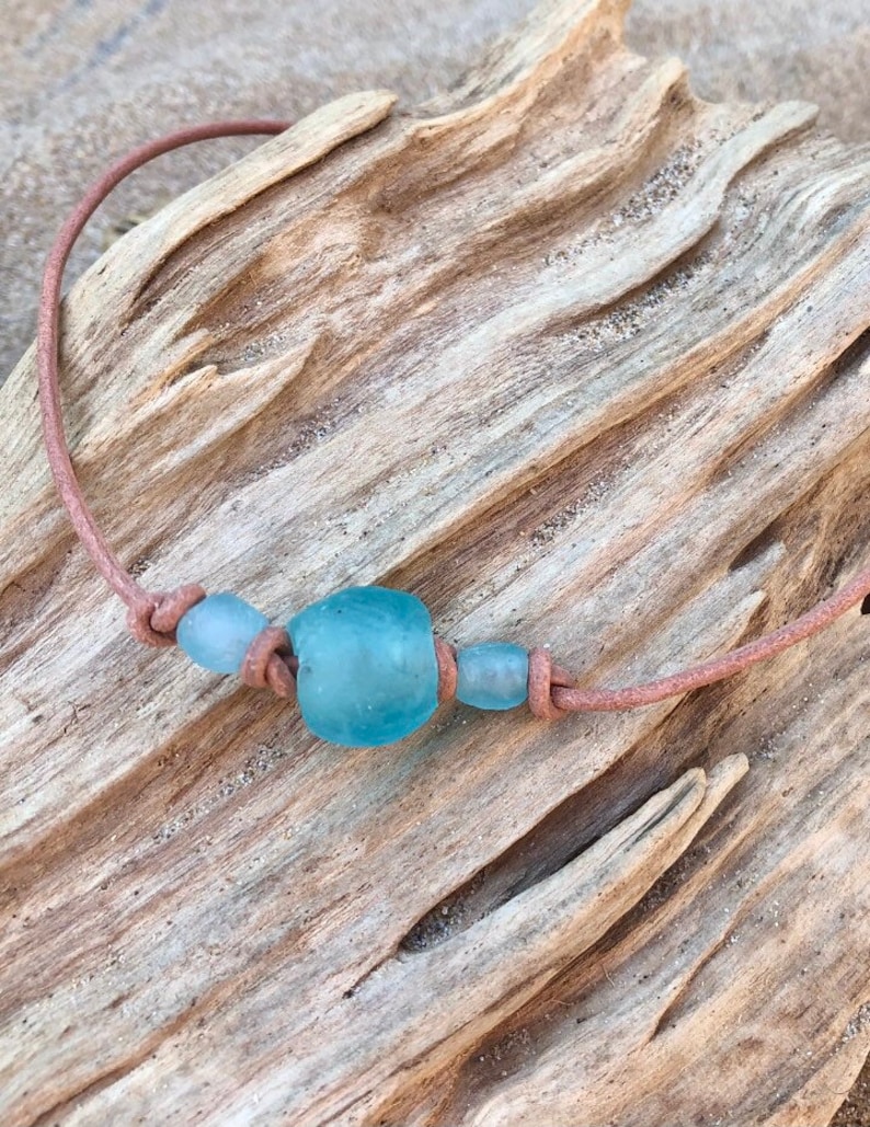 May include: A light blue glass bead necklace with a brown leather cord. The necklace is on a piece of driftwood.