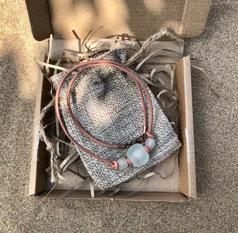May include: A light blue glass bead necklace with brown leather cord, packaged in a brown box with burlap fabric.