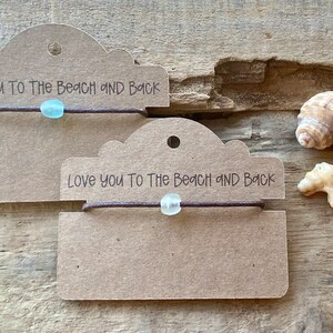 Pode incluir: Duas pulseiras com uma conta azul clara e um cord&atilde;o marrom s&atilde;o exibidas em etiquetas marrons. As etiquetas dizem "Love you to the beach and back". Conchas marinhas ao lado das pulseiras em uma superf&iacute;cie de madeira.