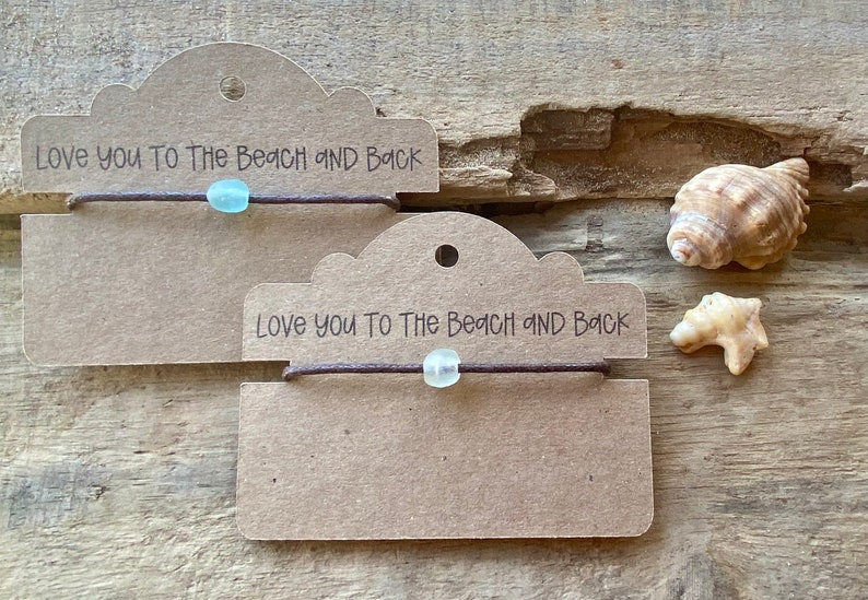 Pode incluir: Duas pulseiras com uma conta azul clara e um cord&atilde;o marrom s&atilde;o exibidas em etiquetas marrons. As etiquetas dizem "Love you to the beach and back". Conchas marinhas ao lado das pulseiras em uma superf&iacute;cie de madeira.