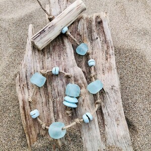 May include: A handmade necklace featuring sea glass beads in shades of blue and white, strung on a natural twine cord. The necklace is displayed on a piece of driftwood, with a matching wooden pendant. The background is sand.