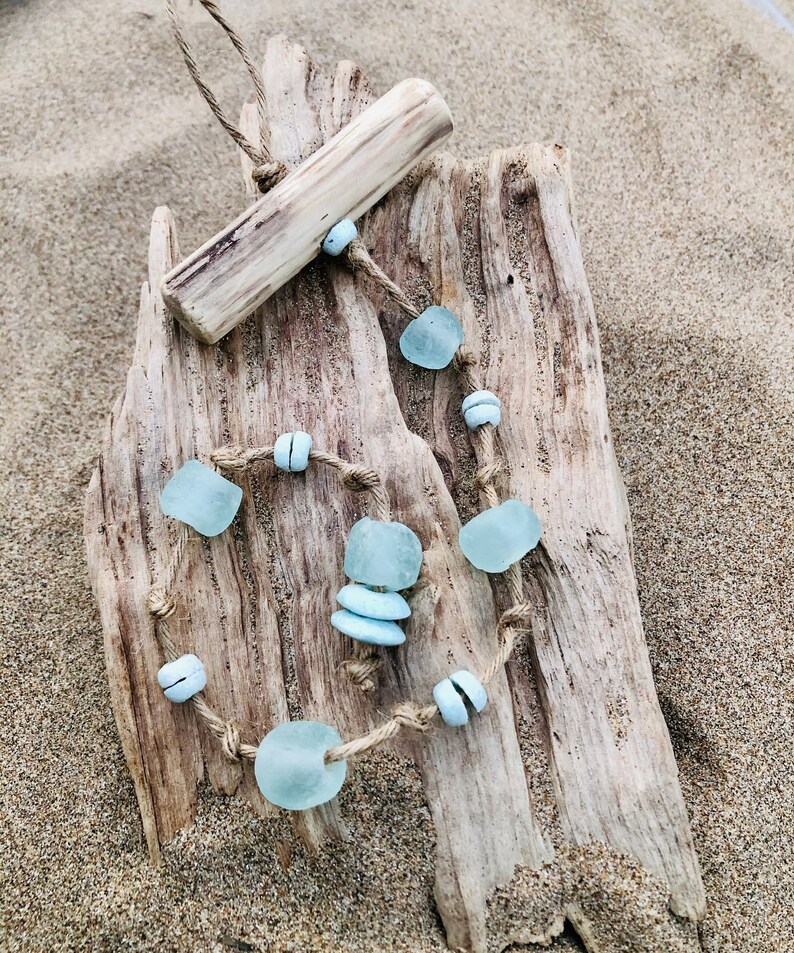 May include: A handmade necklace featuring sea glass beads in shades of blue and white, strung on a natural twine cord. The necklace is displayed on a piece of driftwood, with a matching wooden pendant. The background is sand.