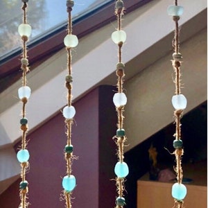 May include: Four strands of decorative hanging beads. Each strand features a series of clear, green, and blue glass beads strung on a natural fiber cord. The beads vary in size and shape, creating a visually appealing, handcrafted look.