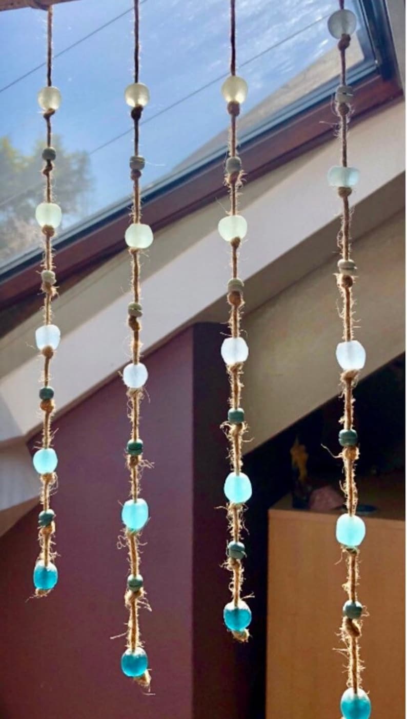 May include: Four strands of decorative hanging beads. Each strand features a series of clear, green, and blue glass beads strung on a natural fiber cord. The beads vary in size and shape, creating a visually appealing, handcrafted look.