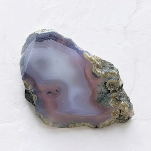 Puede incluir: Una rebanada de ágata pulida con una forma natural e irregular. La piedra presenta bandas de lavanda, malva y blanco, con un borde exterior gris oscuro y áreas con tonos terrosos texturizados. La apariencia general es suave y translúcida.