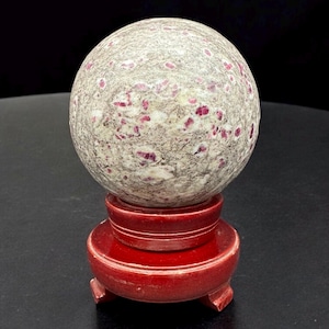 May include: A speckled, light gray sphere with red inclusions rests on a small, round, red wooden stand. The sphere has a textured surface and is the focal point against a dark background. The stand has a tiered design.