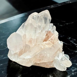 Puede incluir: Un cúmulo de cristales de cuarzo transparentes con un tono rosado. Los cristales están dispuestos en un cúmulo, con algunos de los cristales apuntando hacia arriba y otros hacia abajo. Los cristales son translúcidos y tienen un aspecto vítreo.