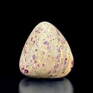 May include: A smooth, egg-shaped stone with a light tan base color, speckled with vibrant purple and magenta inclusions. The stone is set against a black background, highlighting its natural texture and color variations.