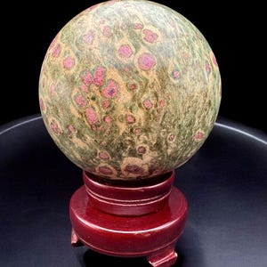 May include: A green and pink sphere with a swirling pattern, resting on a red wooden stand. The sphere is made of a natural stone, possibly jasper or agate.