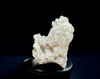 Puede incluir: Un grupo de cristales de cuarzo transparente, con una superficie rugosa y texturizada, se asienta sobre un pedestal negro sobre un fondo oscuro. La formación de cuarzo tiene una estructura cristalina y una forma natural y orgánica.
