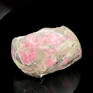 May include: A natural stone with a mix of pink and off-white colors. The stone has a rough, textured surface with patches of vibrant pink crystals embedded within the lighter-colored base. The stone is set against a black background.