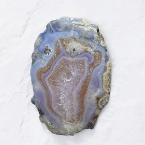 Op de afbeelding: Een gepolijste agaat schijf met een natuurlijke, onregelmatige vorm. De steen heeft concentrische banden van kleur, waaronder tinten van lavendel, bruin en blauw. Het midden heeft een lichtere, doorschijnende tint, waardoor een gelaagd effect ontstaat.