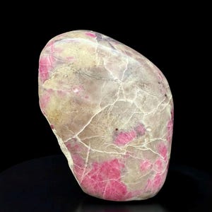 May include: A polished, irregularly shaped stone with a mix of colors. The stone features a base of off-white and gray with vibrant pink patches and veins. The stone is smooth and appears to be a natural mineral specimen.