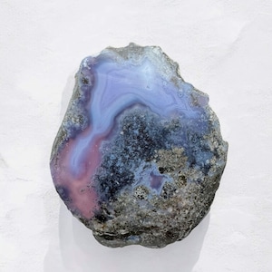 Puede incluir: Una piedra de ágata pulida, de forma irregular, que muestra una mezcla de colores. El centro presenta un tono azul suave, que pasa a rosa y luego a un borde gris oscuro y moteado. La superficie de la piedra tiene patrones intrincados.