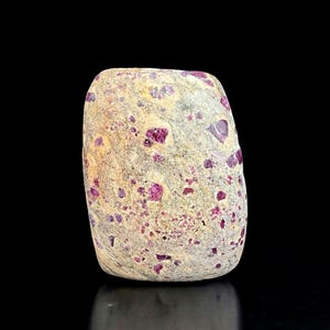 May include: A natural stone with a light beige base color, speckled with numerous vibrant, deep red ruby crystals. The stone has a rounded, oblong shape and a slightly rough texture, showcasing the raw beauty of the gemstones.