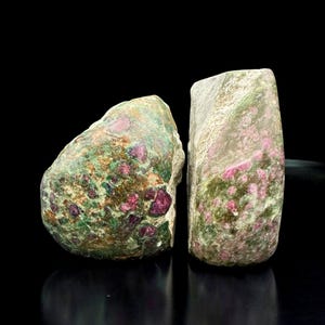 Op de afbeelding: Twee ruwe stenen met een mix van groene, bruine en roze tinten. De stenen hebben een natuurlijke, organische vorm, met zichtbare robijninsluitsels. De achtergrond is effen zwart.