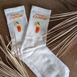 Könnte beinhalten: Ein Paar weiße Socken mit einem orangefarbenen und gelben Motiv eines Cocktailgetränks und dem Text "Power Cocktail" auf jeder Socke.