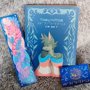 Peut inclure: Un livre bleu avec le titre "TRANSCRIPTEUR 1 AN SOUS T" en texte blanc. Le livre a un motif floral avec une créature à fourrure bleue et rose sur la couverture. Il y a deux signets avec un design similaire. Un signet présente une créature à fourrure rose et bleue et l'autre présente une créature à fourrure violette.