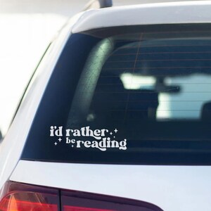 May include: White vinyl sticker on the rear window of a silver car that reads "i'd rather be reading" with stars on either side of the text.