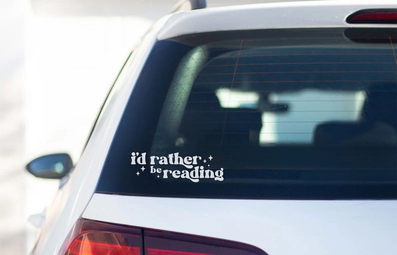 May include: White vinyl sticker on the rear window of a silver car that reads "i'd rather be reading" with stars on either side of the text.