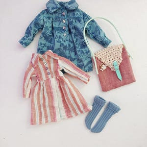 May include: A collection of doll clothes including a blue floral print jacket, a striped dress, a pink purse with a crocheted flap, and a pair of blue socks. The jacket and dress have button closures.