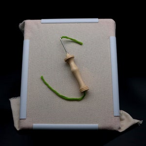 May include: A beige fabric stretched on a white framed square, with a wooden handled punch needle and green yarn. The needle is inserted into the fabric, with the yarn creating a curved line. The background is black.
