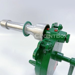 May include: A green metal hand crank sausage stuffer with a silver metal cylinder and a green rubber ring. The stuffer has a handle on the right side and a metal nut on the left side.