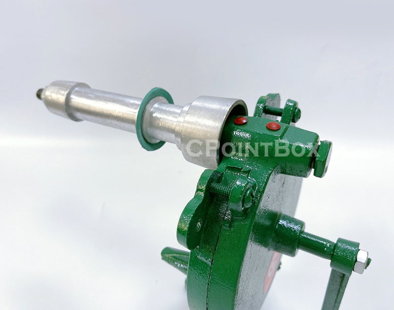 May include: A green metal hand crank sausage stuffer with a silver metal cylinder and a green rubber ring. The stuffer has a handle on the right side and a metal nut on the left side.