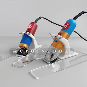 Könnte beinhalten: Zwei elektrische Haarschneider mit unterschiedlich farbigen Gehäusen werden auf durchsichtigen Acrylständern präsentiert. Die Haarschneider sind mit Kabeln verbunden und haben einen roten Knopf an der Seite. Die Ständer sind H-förmig. Der Text "@CPOINTBOX" ist im Bild sichtbar.