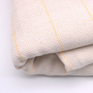 May include: A close-up of a folded piece of off-white fabric with thin yellow stripes. The fabric is woven and has a slightly textured surface.